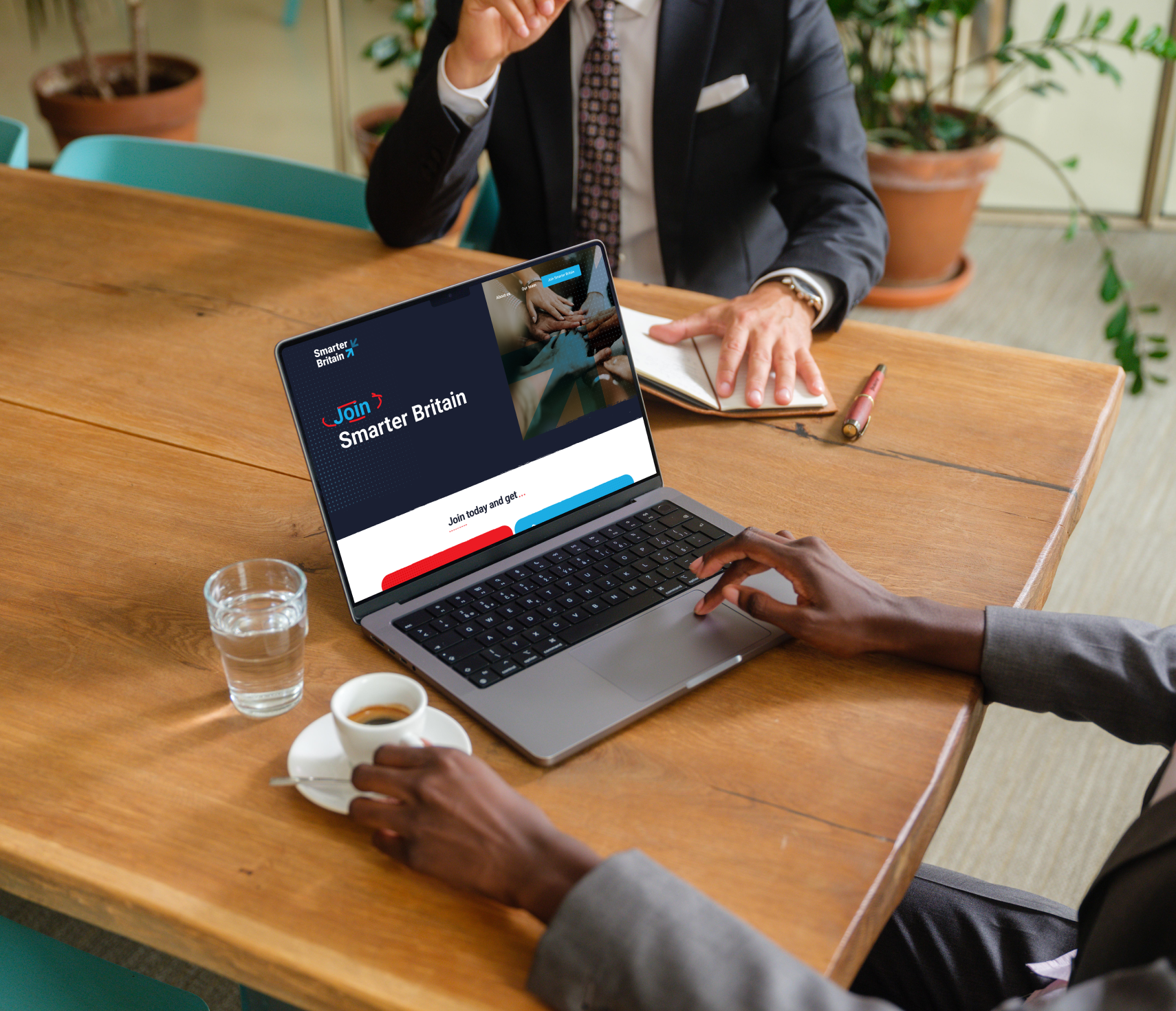 A person sits at a table using a laptop displaying the Smarter Britain website, while another takes notes with a pen. A cup of coffee, glass of water, and potted plant complete the scene.