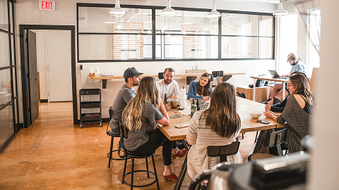 A group of people sit around a wooden table discussing bespoke software development in a modern, bright office space, while another person works alone on a laptop near large windows and glass doors.