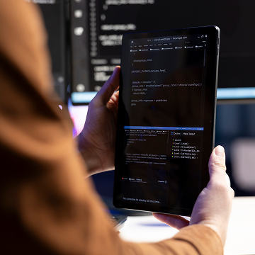 A person holds a tablet displaying computer code, with a desktop monitor showing more code in the background. The scene suggests programming or software development.