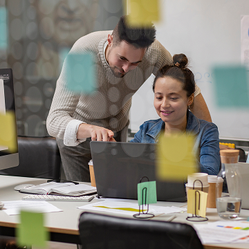 Two people work together at a desk covered with papers, coffee cups, and office supplies. One person sits at a laptop while the other stands beside, pointing at the screen. Colorful sticky notes are visible on glass in the foreground.