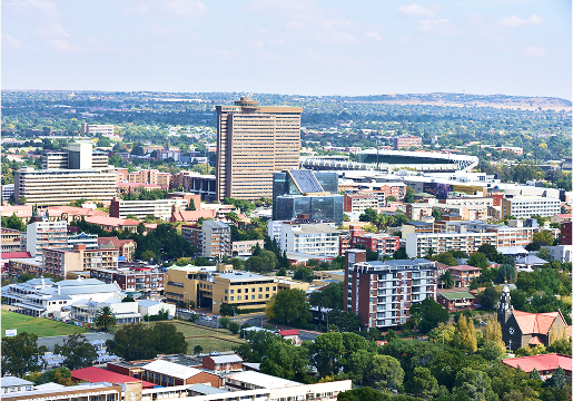 Aerial view of a cityscape with numerous mid-rise buildings, green trees, and a prominent tall building in the center, under a clear blue sky.