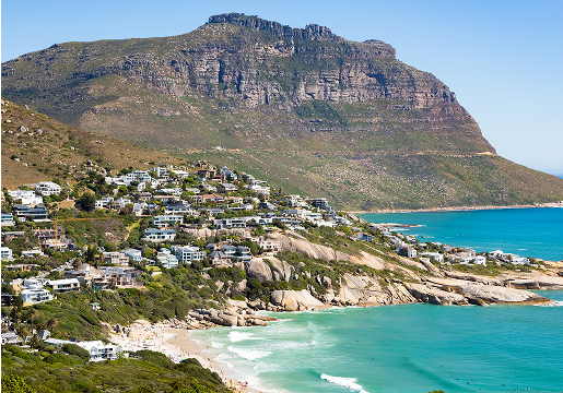 Coastal town with white houses on green hillsides overlooking turquoise ocean water and sandy beaches, with a large rocky mountain in the background under a clear blue sky.