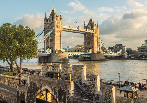 Tower Bridge in London spans the River Thames at sunset, with its two iconic towers and bascules raised. People are walking and relaxing by the riverside, and historic stone walls are visible in the foreground.