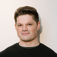A man with short brown hair wearing a black crewneck shirt stands against a plain, light-colored wall, looking at the camera with a neutral expression.