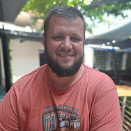 A man with a beard, smiling and wearing a red graphic t-shirt, sits outdoors at a table. Greenery and shaded areas are visible in the background on a sunny day.