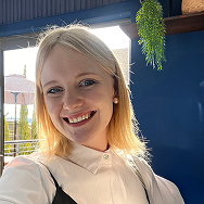 A young woman with blonde hair smiles at the camera indoors. She is wearing a white collared shirt and black suspenders. Sunlight shines in from a window behind her, and a green plant hangs on a blue wall.
