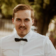 A man with light brown hair and a mustache, wearing a white dress shirt and a black bow tie, stands outdoors with trees and a blurred background.