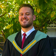 A smiling person in a graduation gown with green and yellow trim, wearing a white shirt and red tie, stands outdoors in front of leafy green trees.