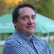 A man with short brown hair wearing a blue and white checkered shirt stands outdoors, with green tents and trees blurred in the background.