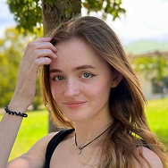 A young woman with long light brown hair, wearing a black top and necklace, stands outdoors by a tree, smiling softly and touching her hair. Green grass and blurred foliage are visible in the background.
