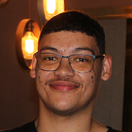 A young person with short, dark hair and glasses smiles at the camera. Warm, exposed light bulbs hang on the wall in the softly lit background.
