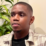 A young man with short hair and an earring, wearing a plaid shirt over a black top, sits indoors with green plants and a light-colored wall in the background.