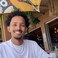 A smiling man with short curly hair and a goatee sits at an outdoor café, wearing a white shirt and gold necklace. A colorful umbrella and hanging plants are visible in the background.