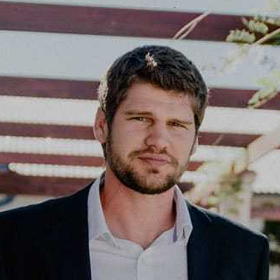 A man with short brown hair and a beard, wearing a white shirt and dark jacket, stands outdoors with blurred pergola beams and greenery in the background.