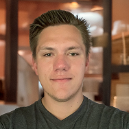 A young man with short, brown hair and a slight smile is standing indoors. He is wearing a dark gray shirt, and the background is softly blurred with warm lighting and wooden elements.