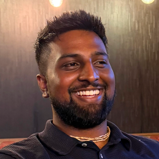 A man with short dark hair and a beard smiles while seated indoors, wearing a black Bitcube collared shirt, a necklace, and an earring. Warm lighting and a dark, textured background are visible behind him.