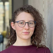 A person with curly brown hair, wearing glasses and a maroon top, stands in front of a light-colored wall, looking at the camera with a neutral expression.
