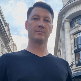 A man wearing a black t-shirt stands outside in a city, with tall, ornate buildings and a partly cloudy sky in the background. The photo is taken from a low angle.
