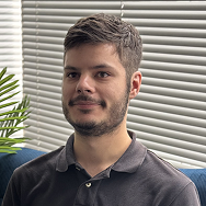 A man with short brown hair and a beard, wearing a dark polo shirt, sits on a blue couch in front of window blinds. A green plant is partially visible to his left.