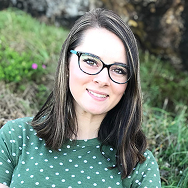 Woman with shoulder-length brown hair and glasses, wearing a green polka dot shirt, smiling outdoors with grass and rocks in the background.