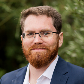 A man with short brown hair, a full reddish beard, and glasses is wearing a navy blue suit and white shirt. He is outdoors with a blurred green background.