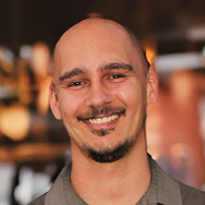 A smiling man with a shaved head, trimmed goatee, and mustache is wearing a gray shirt. The background is softly blurred with warm lighting, suggesting an indoor setting.
