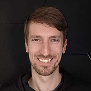 A man with light brown hair and a short beard smiles at the camera. He is wearing a black top and is standing in front of a dark, plain background.