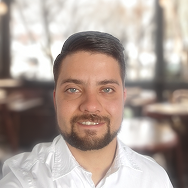A man with short dark hair, a beard, and blue eyes smiles at the camera. He is wearing a white shirt and is standing in a softly lit indoor setting with out-of-focus tables and windows in the background.