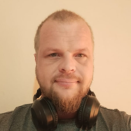 A man with a short beard and mustache is wearing a gray shirt and black headphones around his neck. He has short, light hair and is standing in front of a plain beige background.
