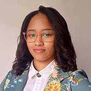 A woman with long dark hair wearing glasses, a white collared shirt, and a green floral blazer, poses in front of a plain light background, looking at the camera with a neutral expression.