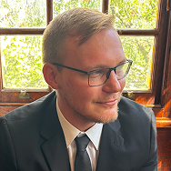 A man with short blond hair, glasses, and a trimmed beard wears a black suit and tie. He is sitting indoors near a sunlit window with a view of green trees outside, looking to his right and smiling slightly.