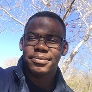A man wearing glasses and a dark jacket smiles outdoors with bare tree branches and a blue sky in the background.
