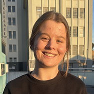 A young woman with light brown hair smiles while standing outside in front of a tall building, bathed in warm sunlight. She is wearing a black shirt.