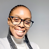 A person with braided hair, wearing black glasses and a white turtleneck, smiles brightly against a plain light background.