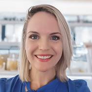A woman with straight blonde hair and bright red lipstick smiles at the camera. She is wearing a blue top and is standing indoors with shelves and jars blurred in the background.