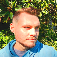 A man with short light brown hair and a trimmed beard, wearing a light blue hoodie, stands outdoors with green foliage and sunlight in the background.