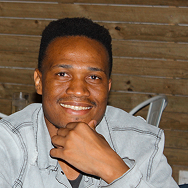 A smiling man sits indoors with his chin resting on his hand. He wears a light denim jacket and is seated in front of a wooden wall.