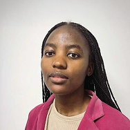 A person with long braided hair, wearing a pink blazer over a beige top, poses against a plain white background and looks calmly at the camera.