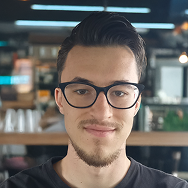 A young man with short dark hair, glasses, and a trimmed beard smiles slightly while standing indoors in a modern, well-lit setting with shelves and plants in the background.