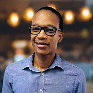 A person wearing glasses and a blue button-up shirt smiles at the camera. The background is softly blurred with warm lights, suggesting an indoor or café setting.