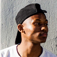 A young man wearing a black baseball cap backwards and a white shirt looks to the side, standing in front of a light-colored textured wall.