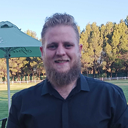 A man with a beard and short hair, wearing a black shirt, smiles outdoors in a park-like setting with trees, grass, and a green umbrella in the background.