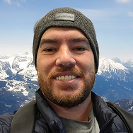A man wearing a gray beanie and a jacket smiles at the camera with snow-covered mountains and a blue sky in the background.