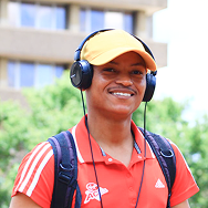 A smiling person wearing a yellow cap, black headphones, a red collared shirt, and a backpack stands outdoors with a blurred building and trees in the background.