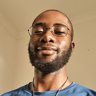 A man with short hair, a beard, and glasses smiles gently while standing indoors in natural light, wearing a blue shirt and a necklace. The background is a plain, light-colored wall.