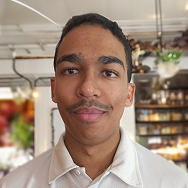 A young man with short dark hair and a mustache, wearing a light-colored collared shirt, stands indoors with shelves and hanging plants in the blurred background.