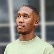 A man with short hair and a trimmed beard, wearing a light green shirt, looks slightly upward with a neutral expression. The background is blurred with soft, grid-like window patterns.