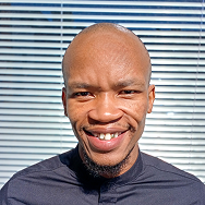 A man with a shaved head and a short beard is smiling at the camera, standing in front of a window with horizontal blinds. He is wearing a black shirt and the lighting is bright.