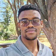 A man wearing glasses and a gray collared shirt stands outdoors near a large tree, with green foliage and sunlight in the background.
