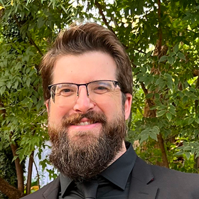 A man with glasses and a full beard, wearing a black suit and tie, smiles in front of leafy green trees outdoors.